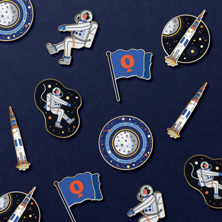 A mix of enamel pins laid out on a blue background. Enamel pins feature the moon, rocket, astronaut, and Questacon 'Q' flag.