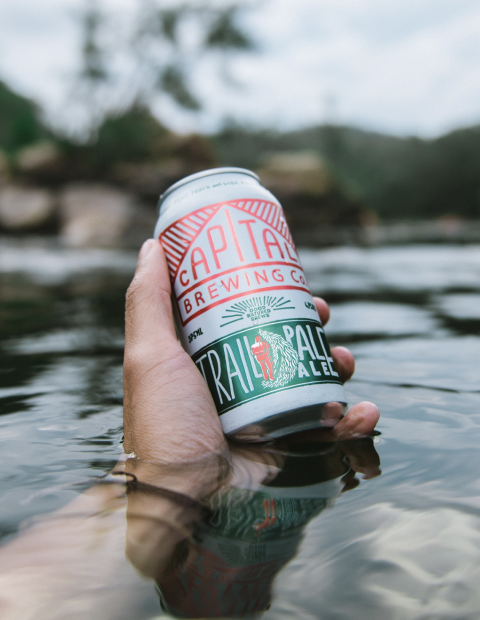 Hand holding a Capital Brewing can with arm submerged in river water.