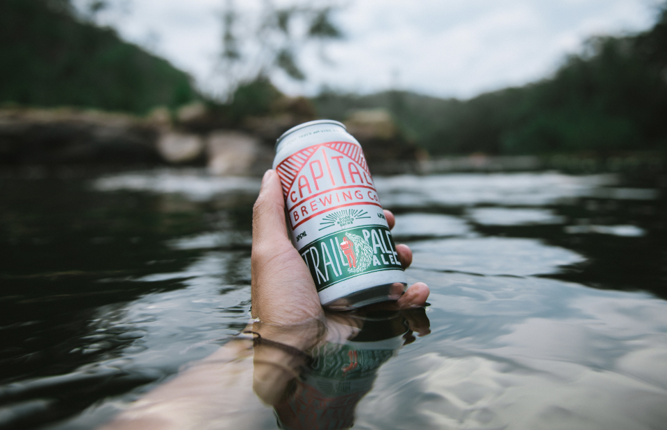 Hand holding a Capital Brewing can with arm submerged in river water.