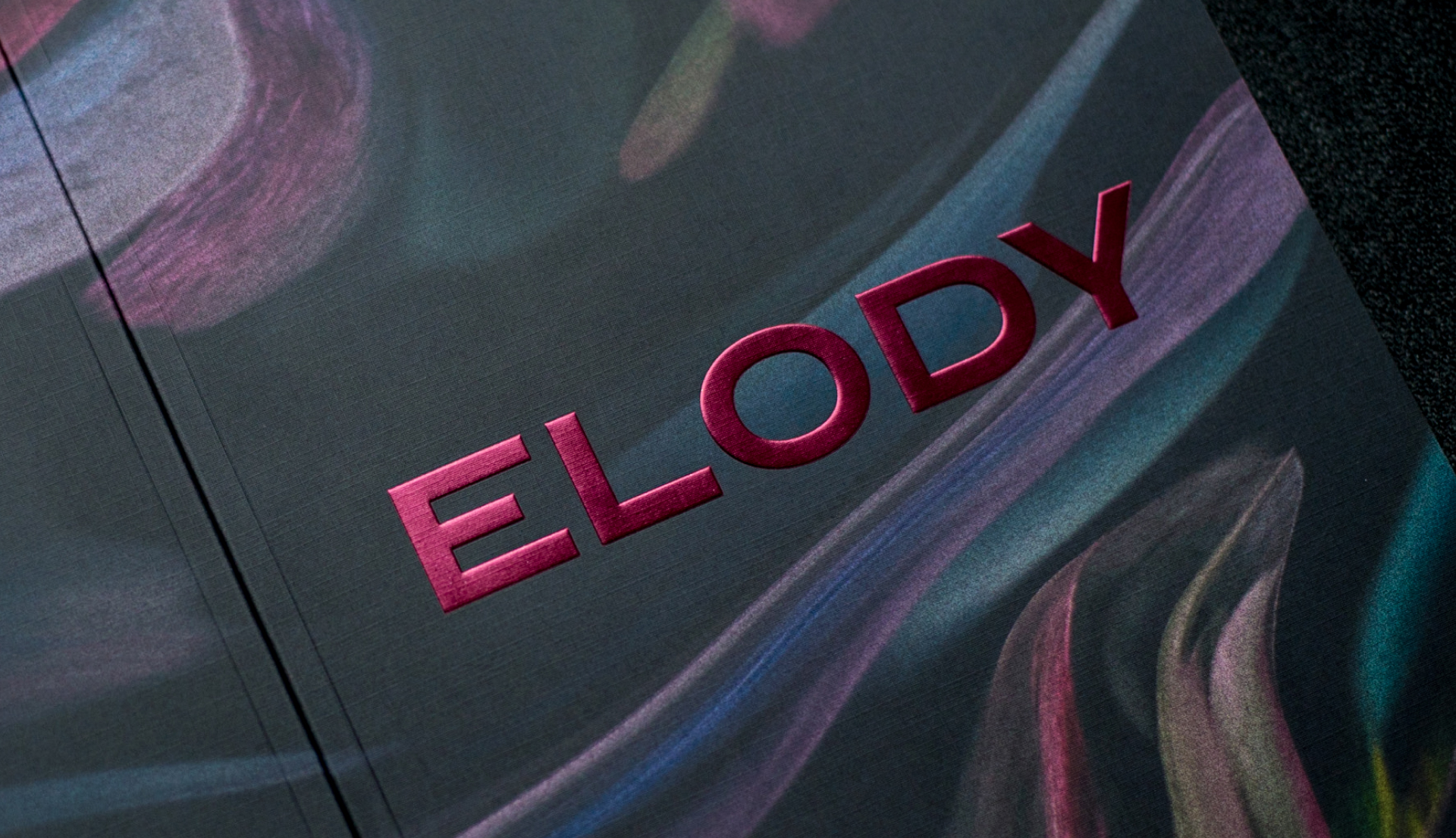 Close up photograph of brochure cover. Elody logo is a maroon foil and embossed. Logo is on a dark and moody abstract background inspired by nature.