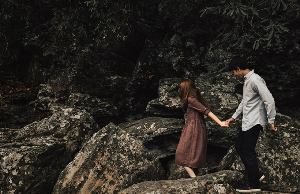 Dark and moody image of couple holding hands and walking over rocks.