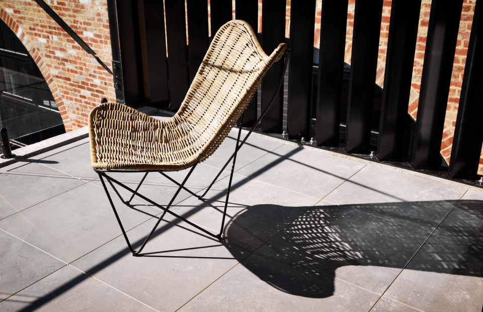 Wicker chair on concrete tiled floor. Harsh sunlight casts a shadow of the chair on the ground.