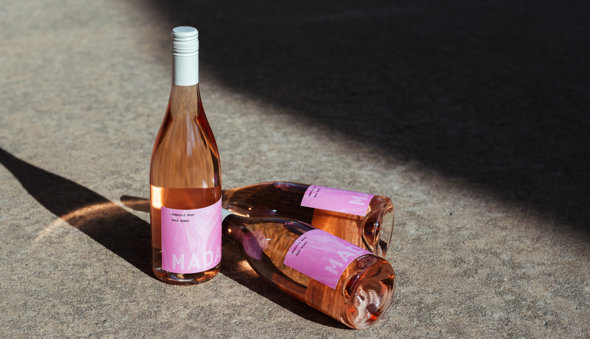 Three rose bottles on concrete ground. Sun is casting long shadows and light is shining through pink glass. Two bottles are lying on their sides, one bottle is standing upright.