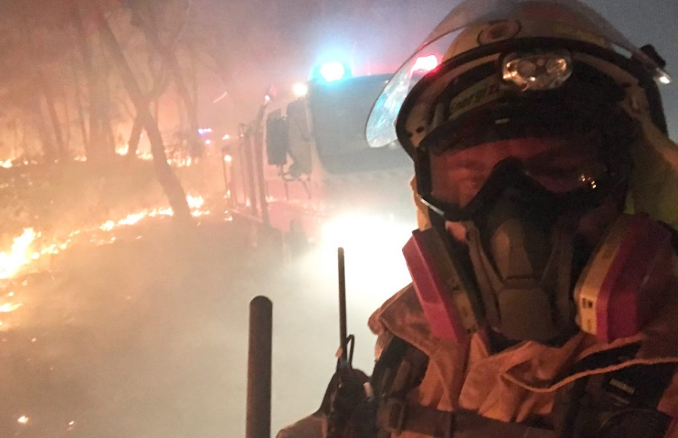 Firefighter wearing full protective gear in the middle of a bushfire. In the background is a firetruck, trees on fire and lots of smoke.