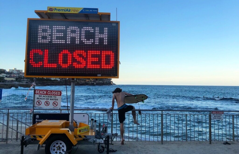 Photo of a surfer with surfboard jumping barrier to get to the ocean. Next to the surfer is an LED sign that reads, "Beach Closed".