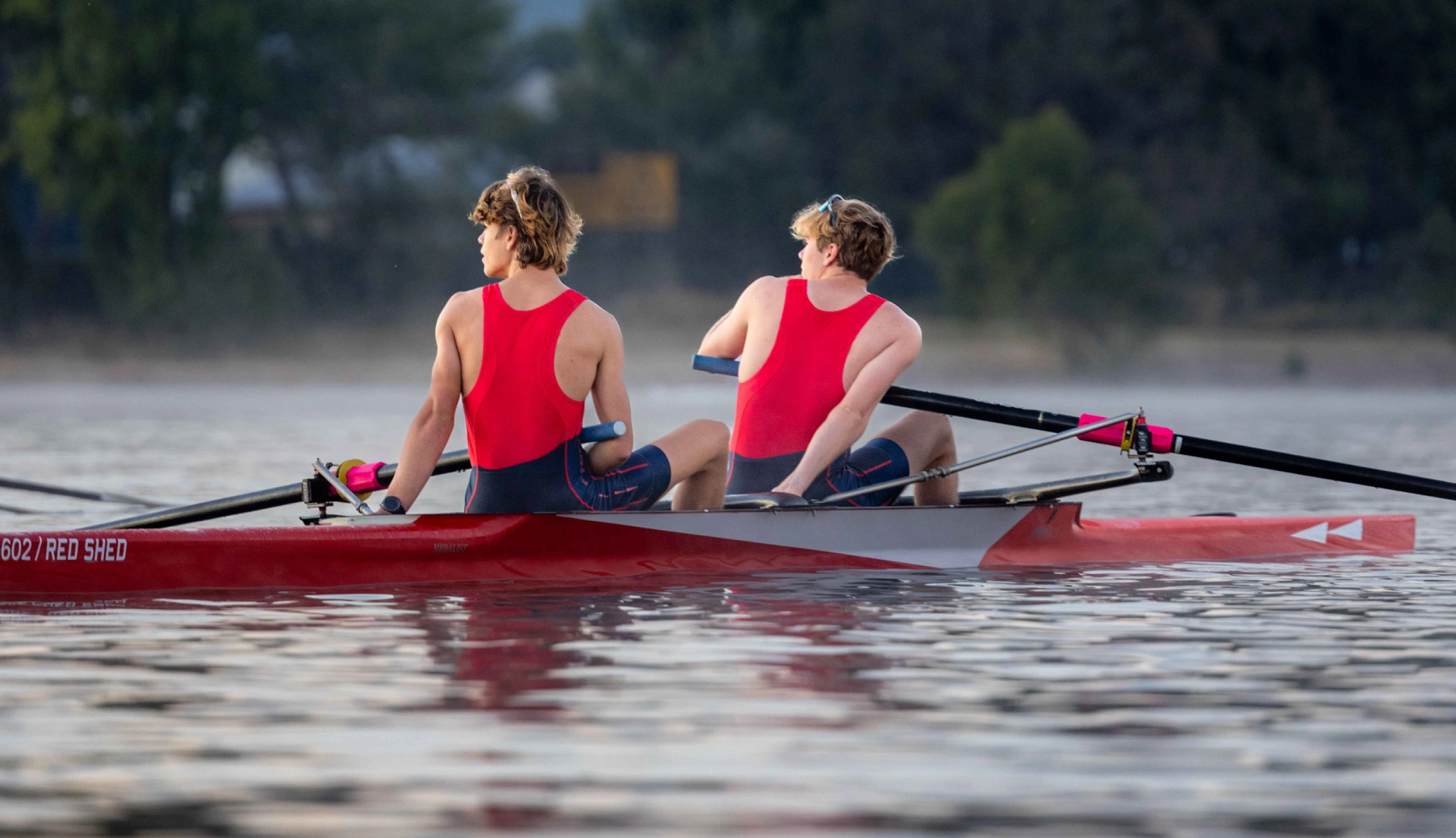 Two rowers in Red Shed row boat on water, wearing Red Shed uniform.